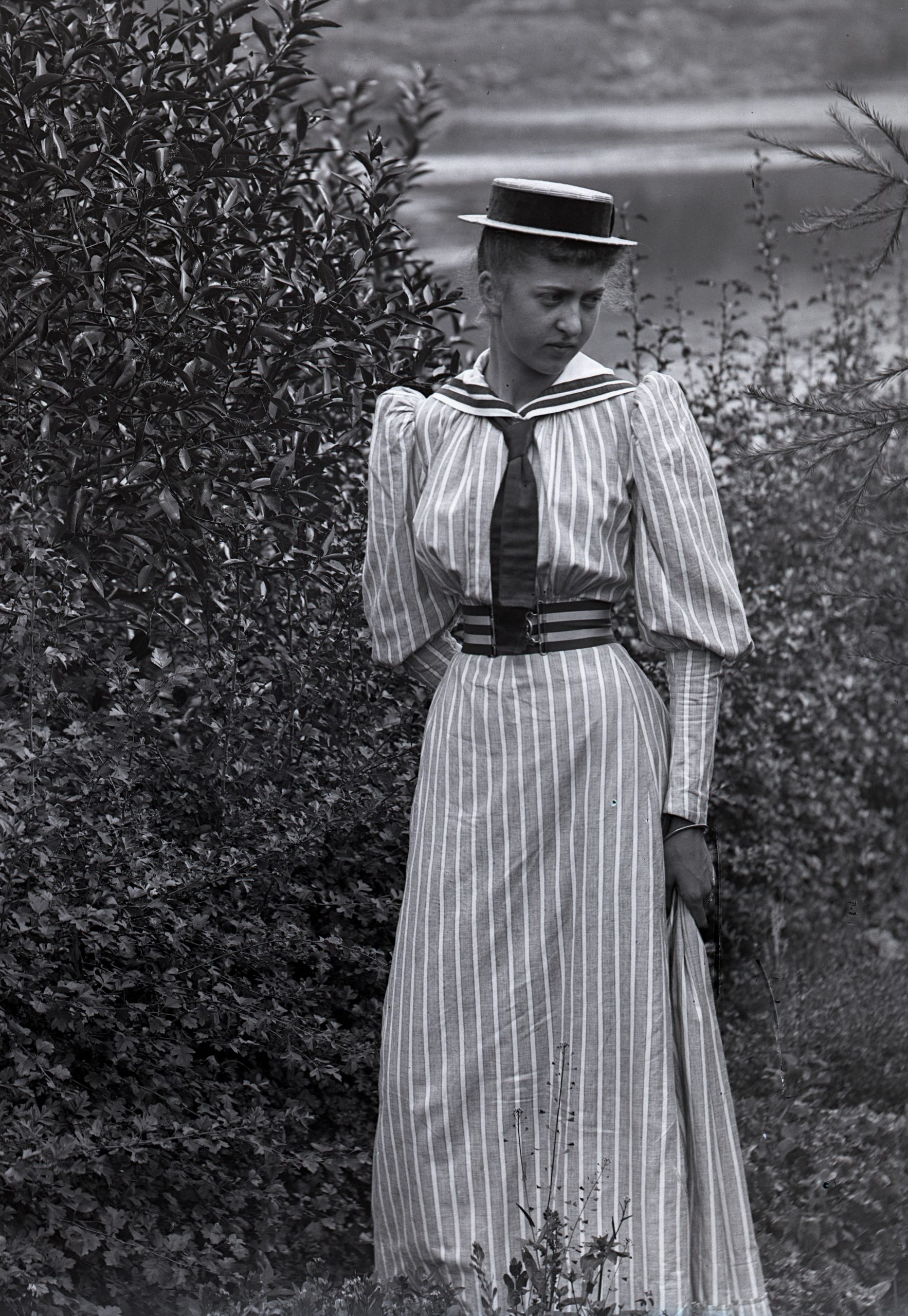 Svartvitt foto. En ung flicka i lång randig dräkt och stråhatt.