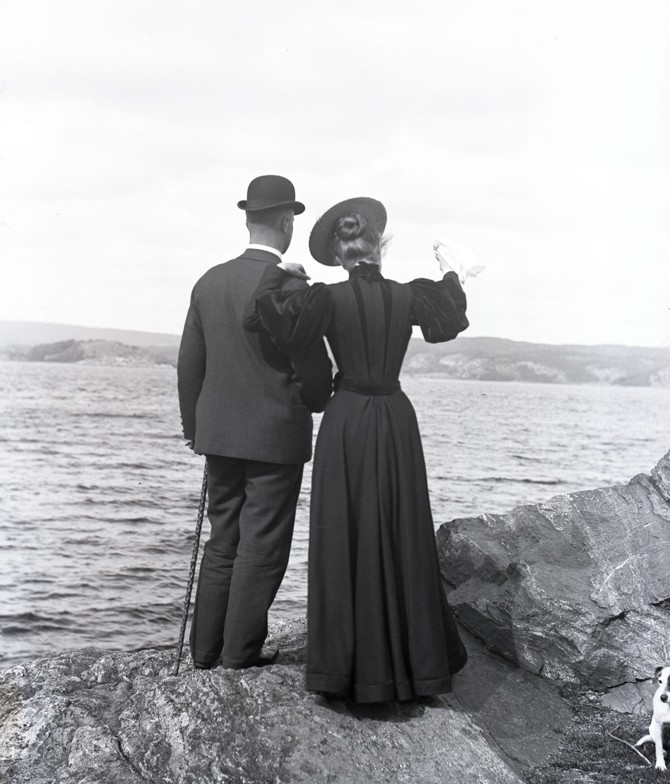 Svartvitt foto. Ett ungt par i mörka dräkter ses bakifrån tittande ut över havet.