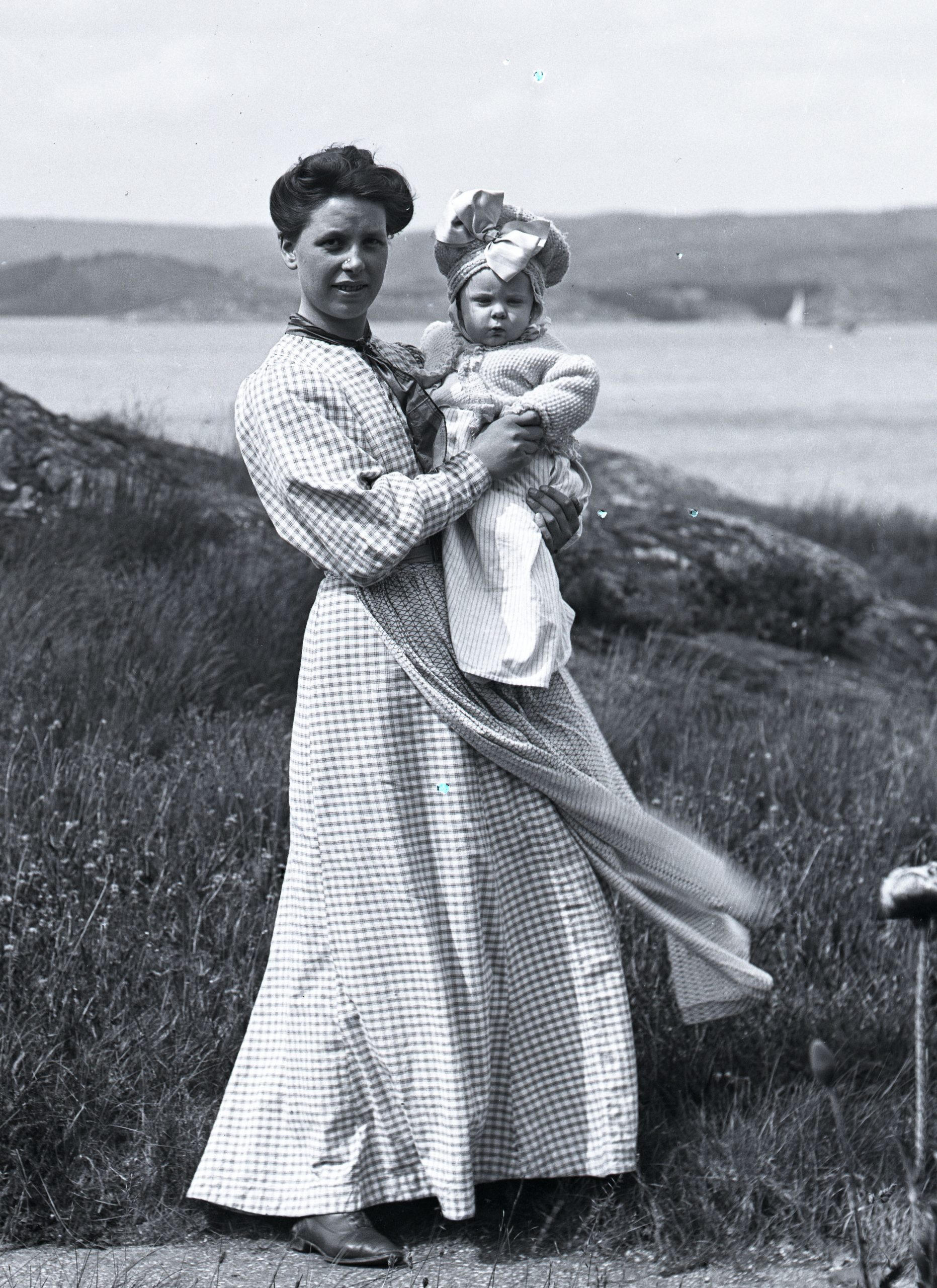 Svartvitt foto. Ung kvinna med baby på berg vid havet.