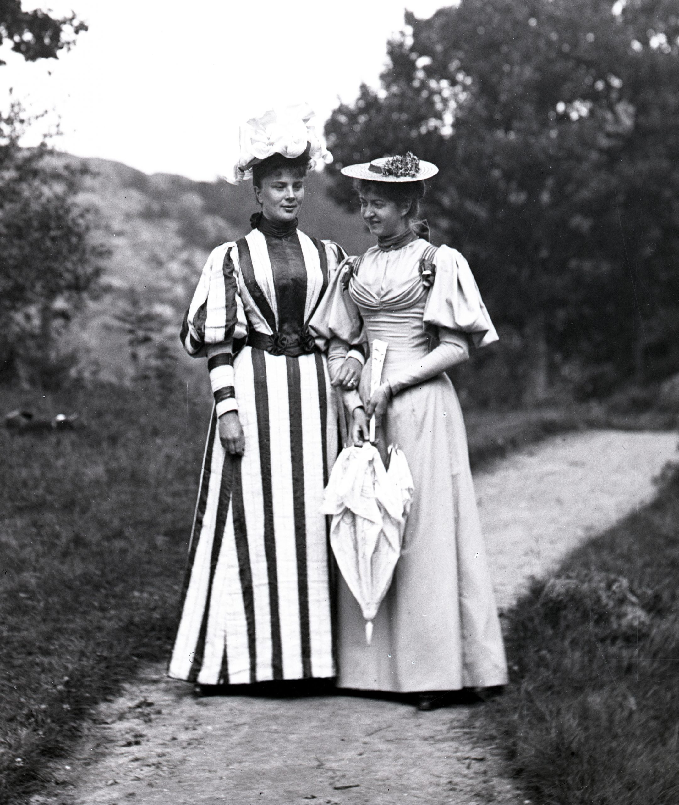 Svartvitt foto. Två unga kvinnor på en promenadväg, båda i lång dräkt och hatt.