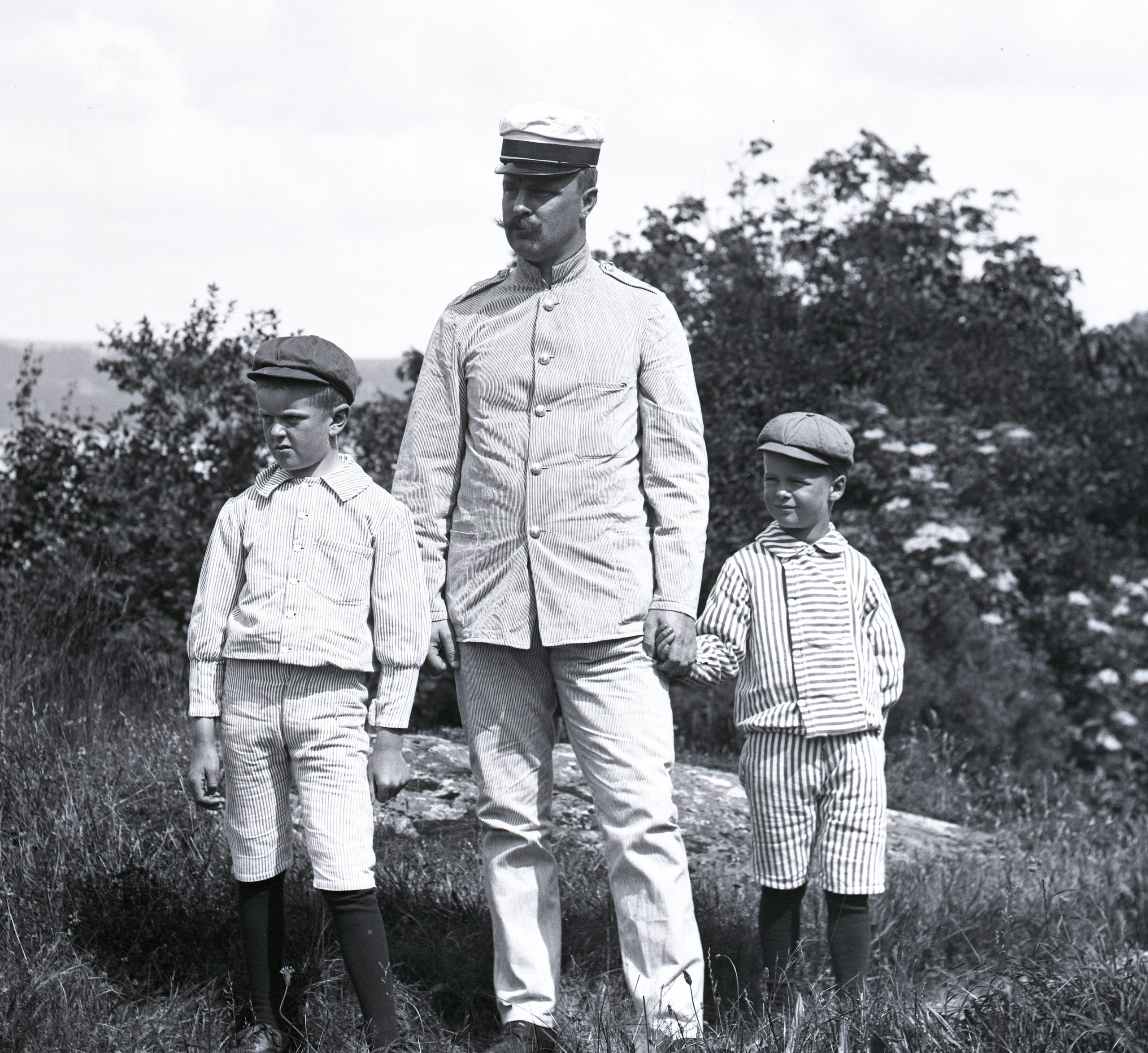 Svartvitt foto. En far i randig kostym och skärmmössa står i trädgården med två söner cirka 6 till 8 år gamla.