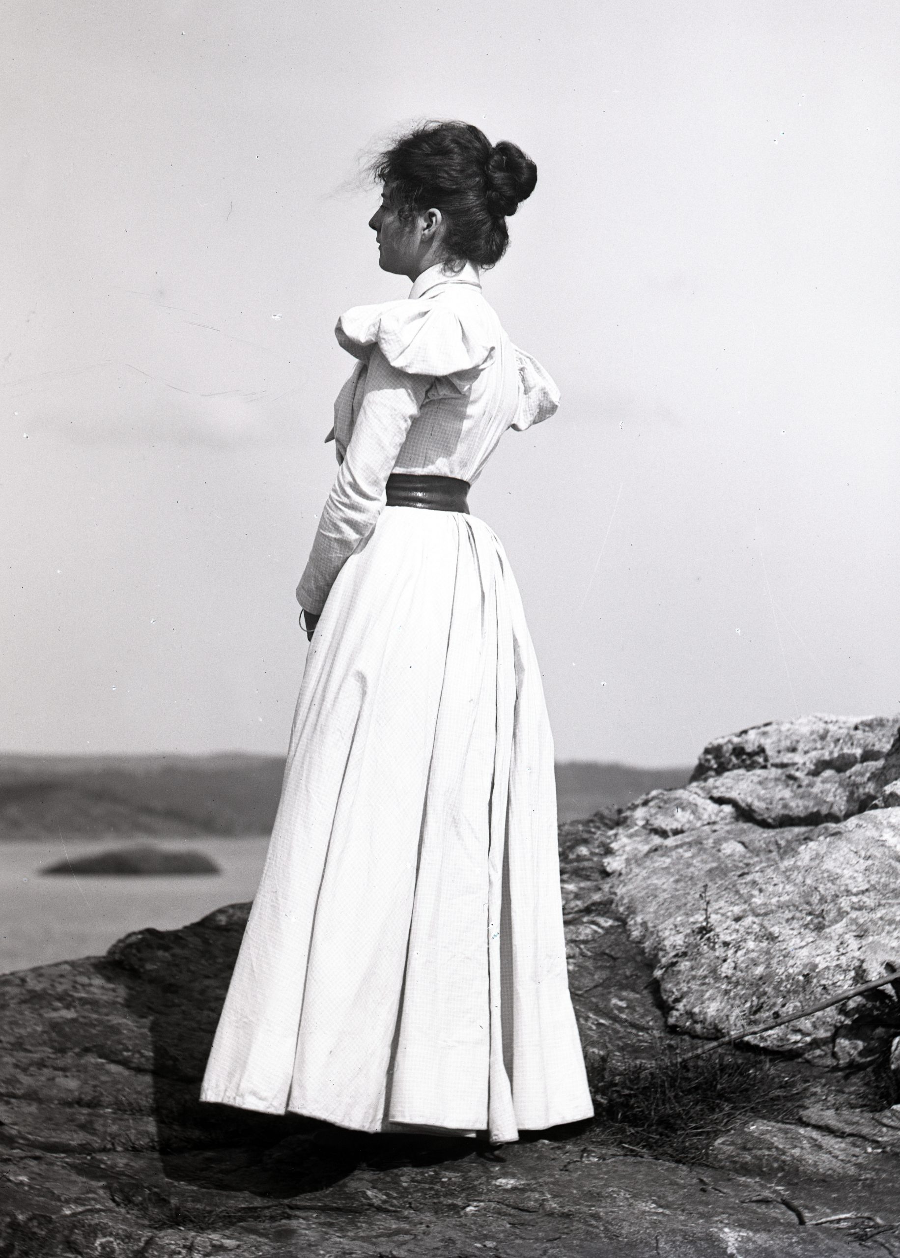 Svartvitt foto. Ung kvinna i lång ljus dräkt står i profil och blickar ut över havet.