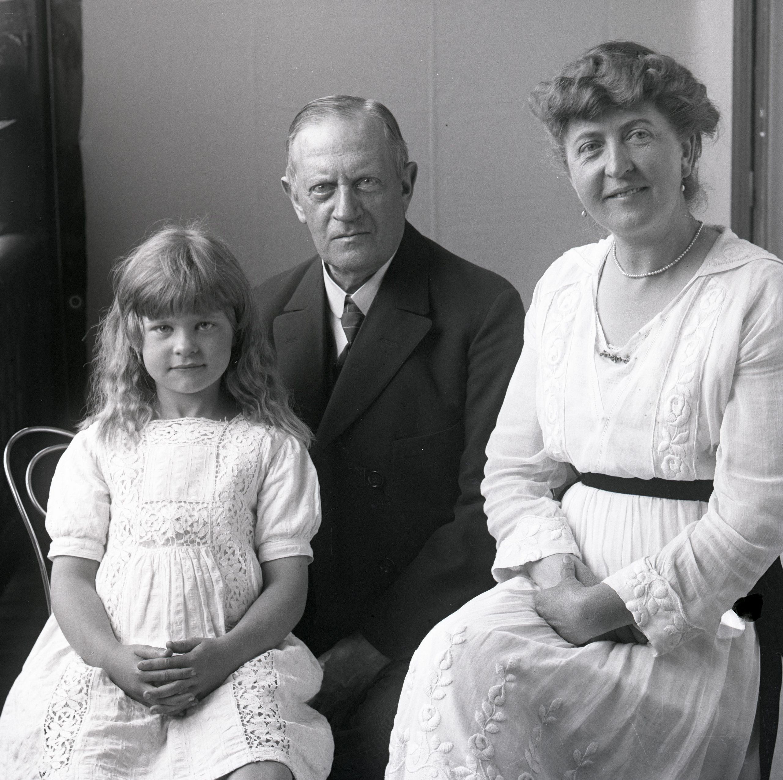Svartvitt foto. En äldre man, en kvinna och en flicka i femårsåldern sitter på en veranda.