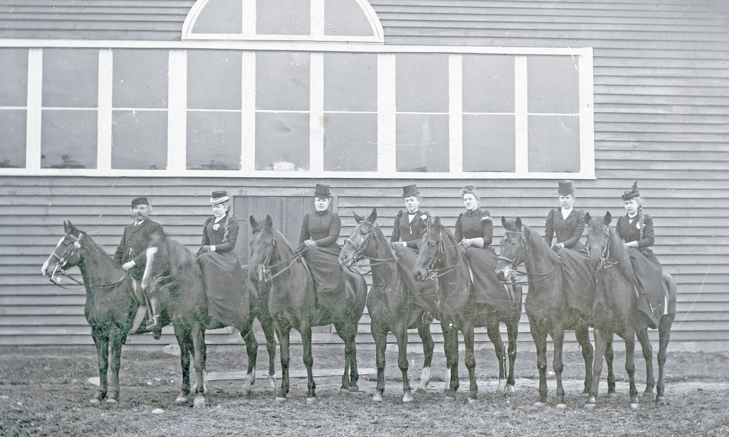 Svartvitt foto. Gruppbild med sju kvinnliga ryttarinnor, klädda i långa kjolar och hatt, sittande till häst framför ett stall.