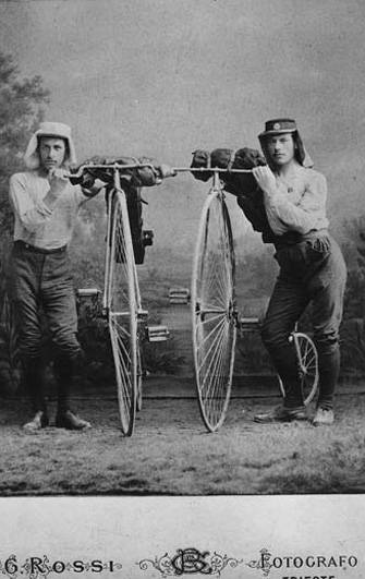 Svartvitt ateljéfoto. Två sportklädda män med skyddande huvudbonader vid sina höghjulingar.