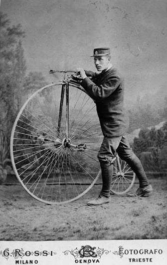Svartvitt ateljéfoto. Man i sportkostym och keps står i profil bredvid sin höghjuling.