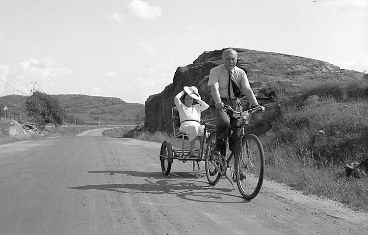 Svartvitt foto. Manlig cyklist kör sittande kvinna i cykelkärra. Hennes hatt flyger nästan av.