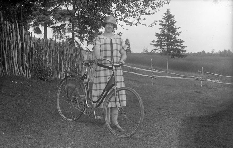 Svartvitt foto. Kvinna i vadlång klänning och rundkullig hatt står vid sin cykel på en gräsbacke.