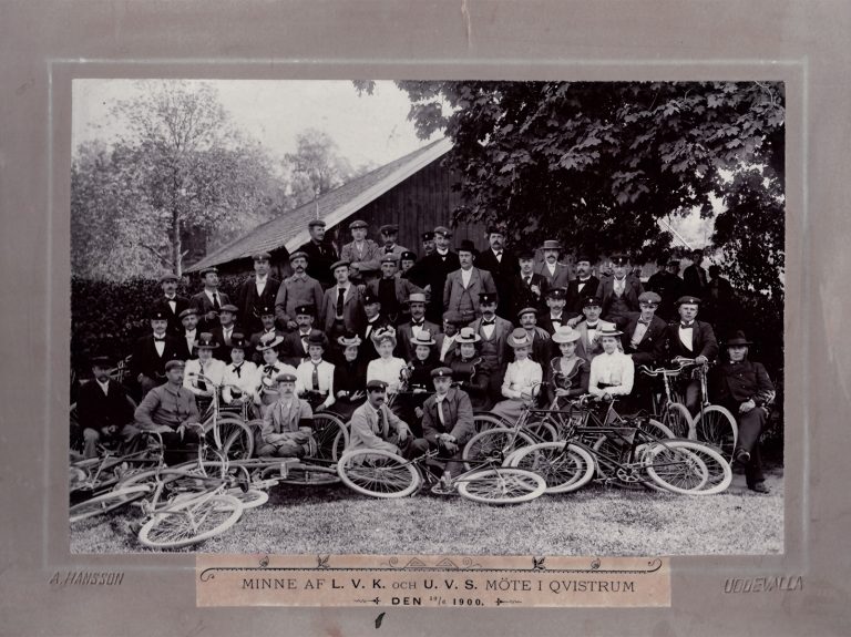 Svartvitt gruppfoto: Ett femtiotal både manliga och kvinnliga cyklister med några liggande cyklar framför sig,
