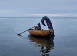 Foto. Person som ror en träeka på hav i stiltje. på ekans högra sida kommer en gigantisk tentakel upp ur havet och reser sig över båten.
