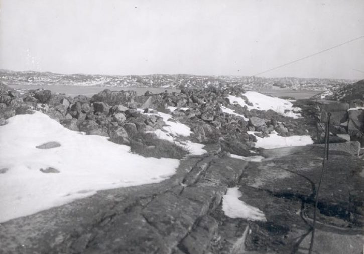 Svartvitt foto. Ett 24 meter långt röse där stenarna har rasat ut åt olika håll. Snödrivor täcker delar av röset. Till vänster finns ett järnstaket.