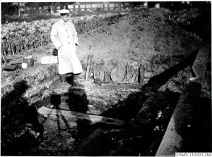 Svartvitt foto: Man i keps och lång rock inspekterar en utgrävning. Trärester skymtar i jorden.