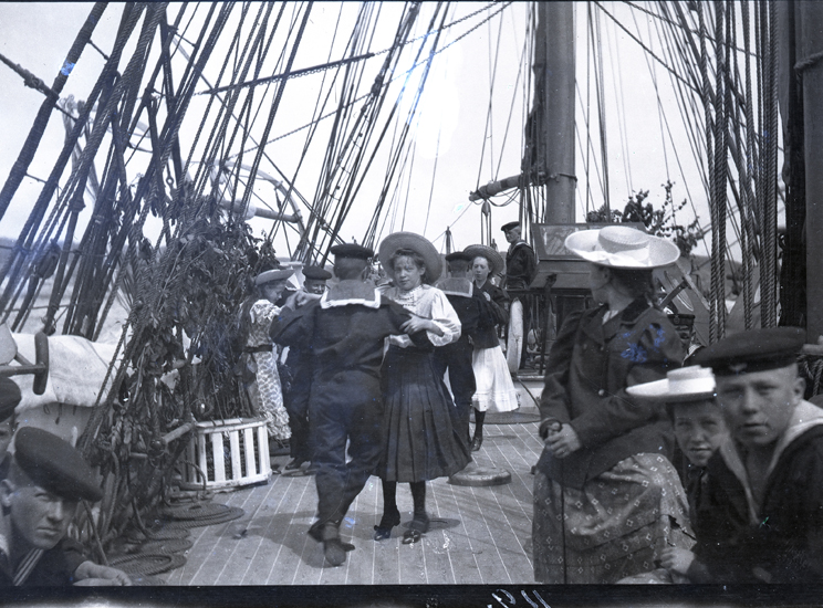 Svartvitt foto: Tre unga par, pojkar i matroskläder och flickor med vadlånga kjolar och stråhattar, dansar ombord på ett lövprytt fartyg.