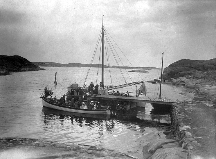 Svartvitt foto. Utmed en brygga ligger tre delvis lövprydda båtar. Ett tjugotal finklädda kvinnor och män kliver ombord.