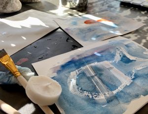 Foto. Pysselbord. Närbild på pensel, ett stearinljus och akvarellpapper med blå färg på och en stiliserad fisk.