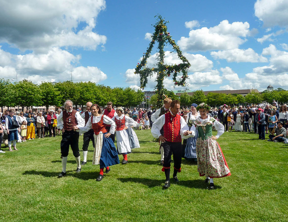 Foto: Traditionellt midsommarfirande på en gröngräsmatta. I Bakgrunden en midsommarstång och folksamling. I förgrunden dansar fyra stycken medelålders par gammeldans i folkdräkter.
