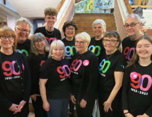 Foto. 11 personer med svarta t-shirtar med Riksteaterns 90-års logga står under framför en trappa i Bohusläns museum. Loggan är i klar rosa eller grön färg. Alla personerna ler och tittar in i kameran.
