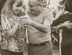 Svartvitt foto. En kvinna med långt hår och stort leende dansar med en man med stort leende och solglasögon. De håller händerna och båda är i övre medelåldern. I bakgrunden ett annat dansande par i oskärpa.