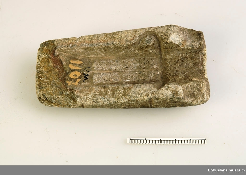 Foto. Ett avlångt liggande  föremål i ljusgrå sten. Stenen är urgröpt till formen av en bronsyxa. En skalstock visar att föremålet är 11,5 centimeter långt och 5,8 centimeter brett.