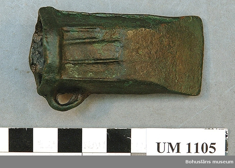 Foto. Avlång grönärgad bronsyxa. En skalstock visar att föremålet är 7 centimeter långt och 3,9 centimeter brett.