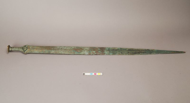 Foto till vänster. Ett grönt ärgat svärd med spetsen åt höger.