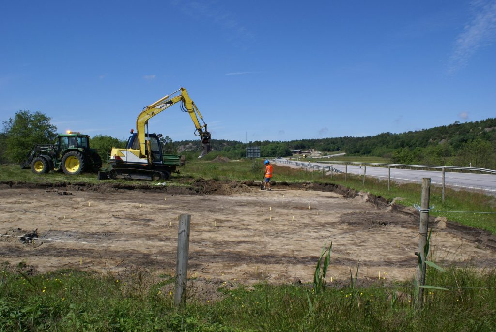 Foto. En kvinna i orange väst står framför en gul grävmaskin. Runtomkring henne syns mörka fläckar i den öppna jorden. Till höger löper en motorväg.  