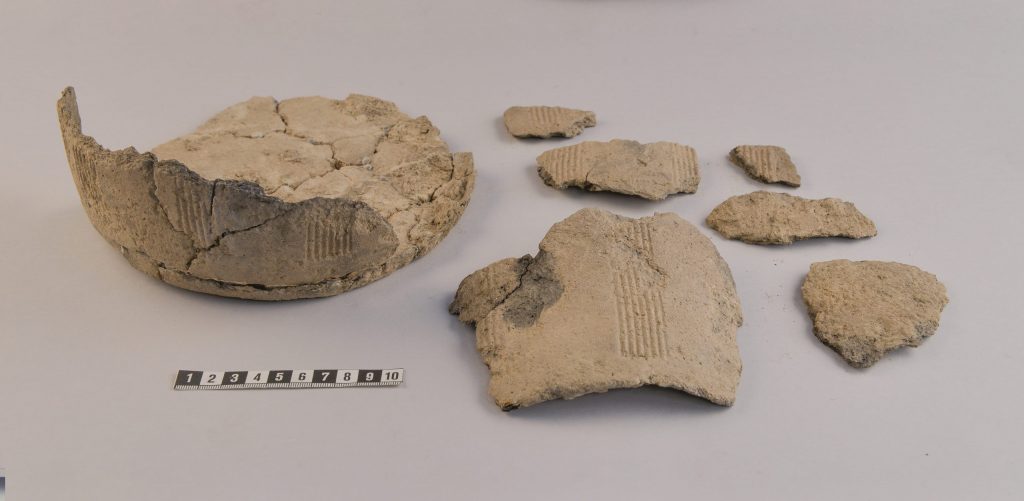 Foto. Sju delar av ett spannformat lerkärl med inristade ränder. En av delarna utgör kärlets botten och är sammanfogad av mindre bitar.