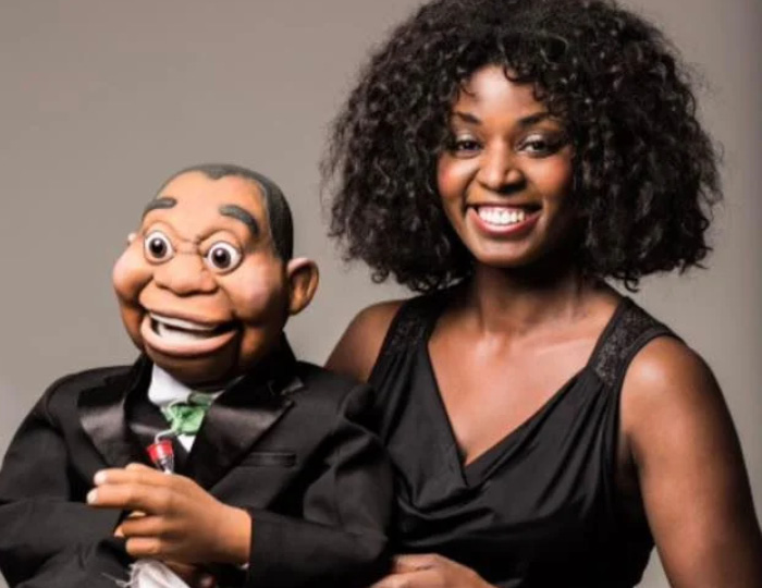 Foto. Caroline Wennergren med sin buktalardocka. Caroline har utsläppt afro och tittar in i kameran och ler, iklädd svart klänning. Dockan är iklädd svart kostym och grön slips, han ler och tittar snett ner till vänster med höjda ögonbryn.