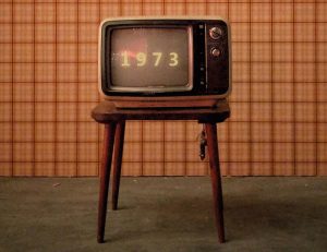 Foto. Retro TV på tik pall i rum med orangerutig tapet och heltäckningsmatta. Siffrorna 1973 står på TV-skärmen.