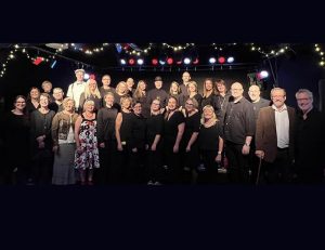 Gruppfoto. Odd Singers står uppställda på en scen med kulörta lyktor. Ett tjugotal personer i medelåldern, blandat män och kvinnor.