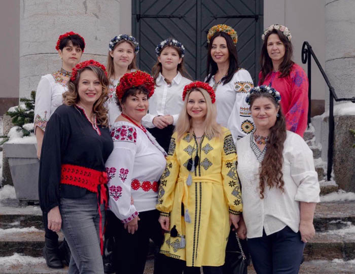 Gruppfoto. 9 st kvinnor i blandade åldrar på en trappa med traditionella blomster huvudsmyckningar.