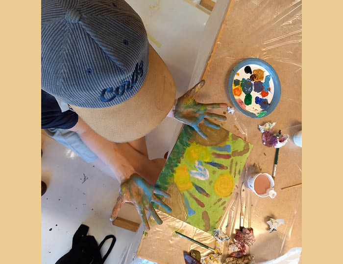 Foto taget ovanifrpn. Barn med keps håller upp sina händer fulla med färg över ett bord där det ligger en målning, penslar och färger utspritt.