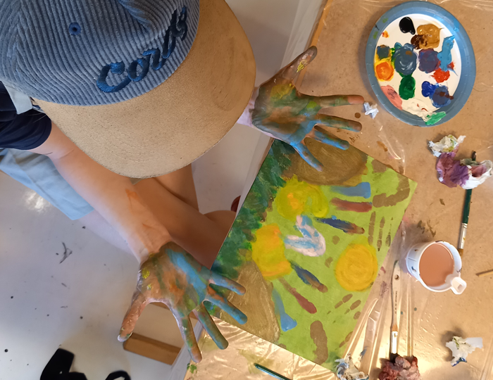 Foto. Ett barn med blå keps och blåfärgade händer leker med färger på ett papper.