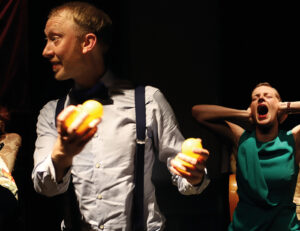 Foto: En man tittar till vänster och ler, med jongleringsbollar i sin hand. I bakgrunden en kvinna som gapar, blundar och håller för öronen.
