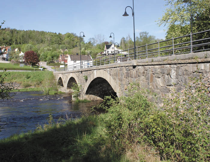 Foto. Kviströmsbro i Munkedal. En stenbro över ett vattendrag. Blå himmel gröna löv på buskar och träd.