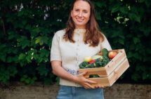 Porträttfoto. Hanna Olvenmark tittar in i kameran och ler. Hon håller en trälåda med grönsaker.