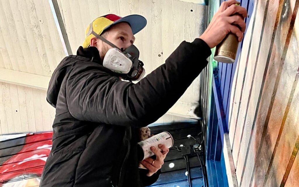 Foto. Ung man i keps och svart jacka med gasmask. målar graffiti på träpanel utomhus.