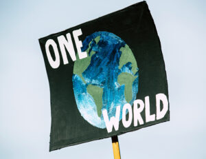 Foto. Demonstrationsplakat mot ljusblå himmel. Svart bakgrund, text i vitt "one World" en målad jordglob i grönt och blått. Plakatet sitter på en träpinne.