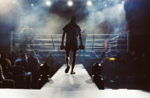 Foto. Boxare som går på catwalk mot boxningsring. Rök och lampor riktade mot ringen.