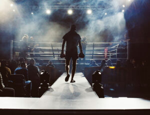 Foto. Boxare som går på catwalk mot boxningsring. Rök och lampor riktade mot ringen.