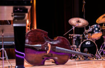 Foto. En scen med instrument. I mitten ligger en cello horisontellt på golvet, till höger en trumma. I förgrunden syns ett ben tillhörande ett klaviatur.