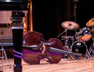 Foto. En scen med instrument. I mitten ligger en cello horisontellt på golvet, till höger en trumma. I förgrunden syns ett ben tillhörande ett klaviatur.