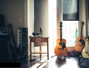 Foto. Rum med instrument uppställda, tre gitarrer till höger en mikrofon i mitten ett elpiano och en bas till vänster. Fönster med klar sol i bakgrunden.