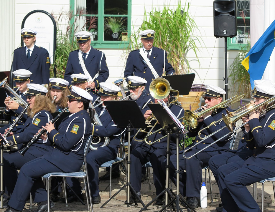 Foto. Uddevalla Stadsmusikkår utomhus. Ett tio tal musikanter som sitter och står och spelar musik,