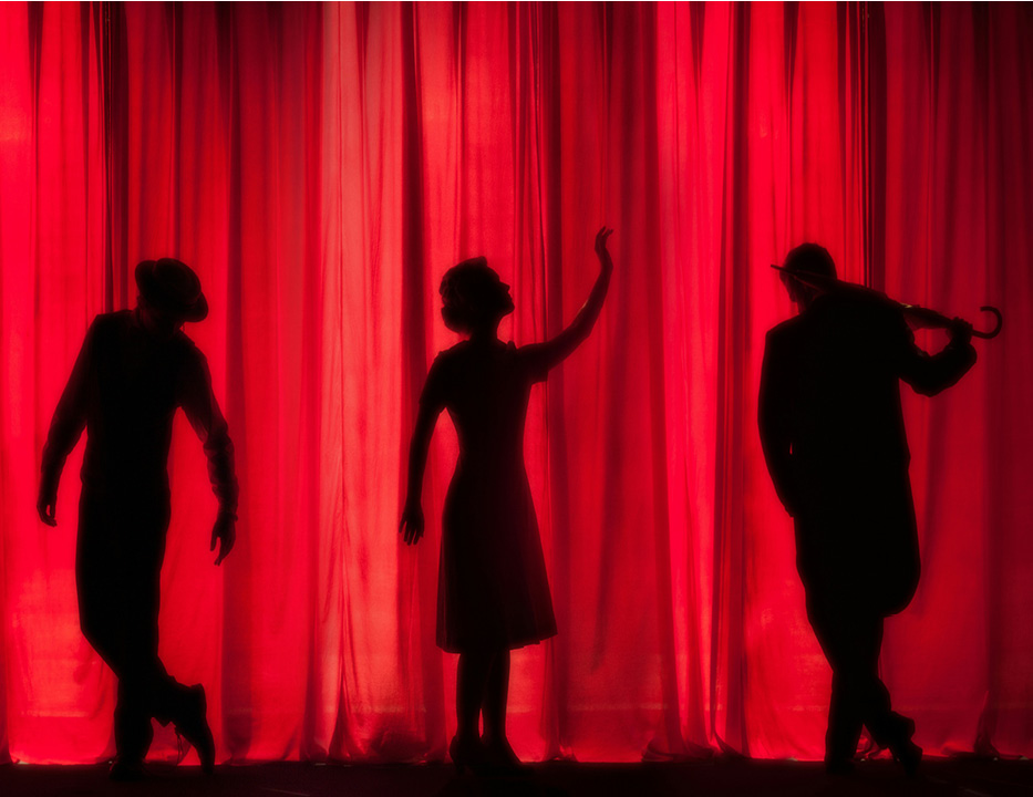 Foto en röd ridå med siluetter av tre människor, Till vänster person med hatt, mitten person i klänning med blicken riktad mot sin hand som hålls upp i skyn, till höger person som spelar på en fiol.