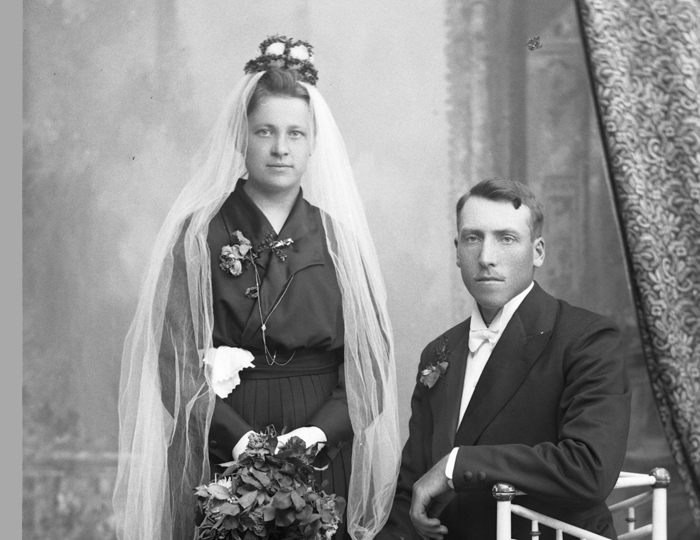 Svartvitt foto. Bruden i svart dräkt och vit slöja står bredvid brudgummen i kostym som sitter i en stol.