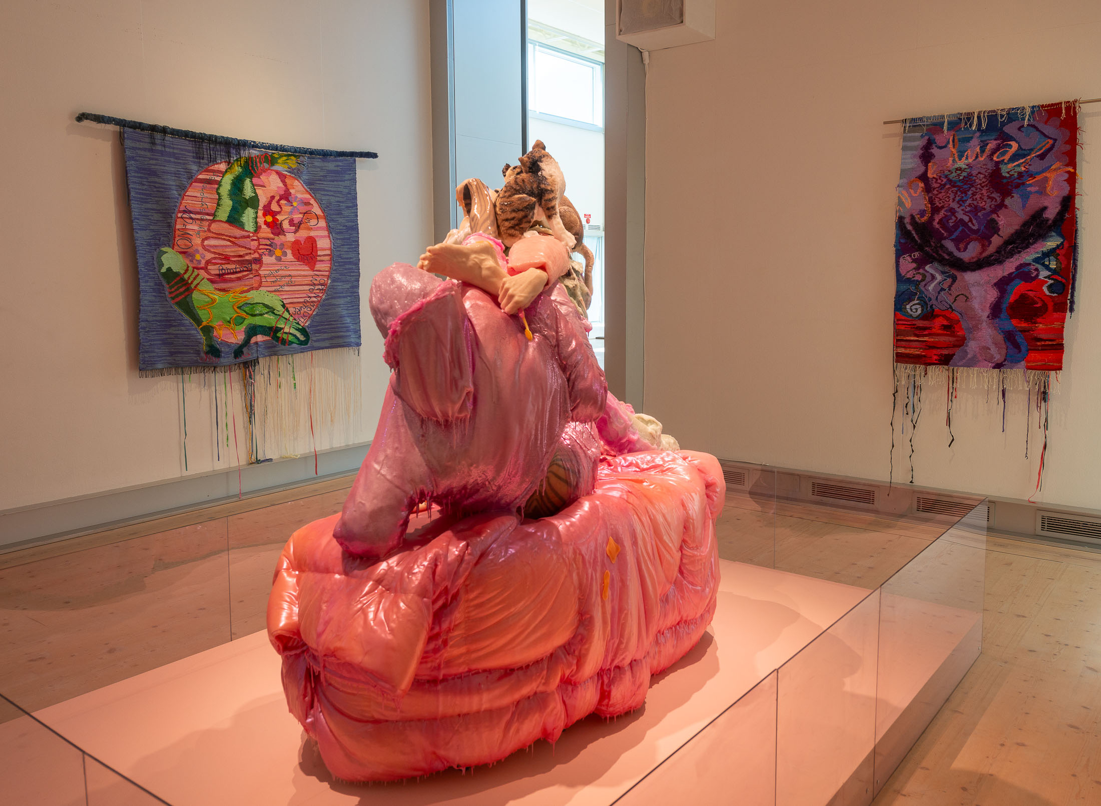 Bilden är tagen i konsthallen på Bohusläns museum. I förgrunden en skulptur som föreställer två kvinnor klädda i rosa. I bakgrunden två vävda konstverk. Rummet har trägolv och vita väggar.