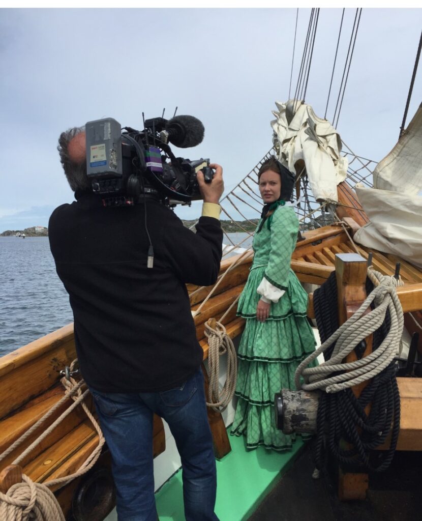 Foto. En man filmar en kvinna klädd i grön 1800-tals dräkt som står i en segelbåt.