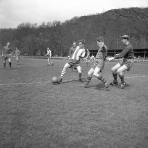 Svartvitt foto. En fotbollsmatch. En ung man i randigt försöker dribbla sig förbi två spelare i helfärgade tröjor, En av försvararna har en keps.