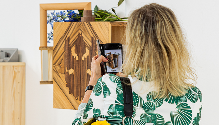 Foto. En blond kvinna fotograferar ett konsthantverk i trä.